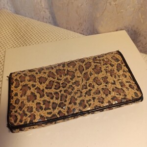 May include: A rectangular clutch wallet with a leopard print pattern in shades of brown and tan. The wallet has a black border and a zipper closure. The wallet is made of a textured material.