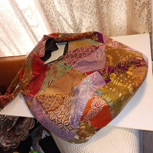 May include: A patchwork tote bag with a variety of patterned fabrics in shades of brown, red, purple, and gold. The bag has a black interior and a long strap. The bag is made of multiple fabric pieces sewn together.