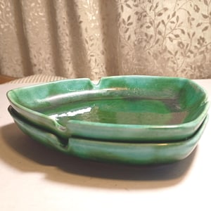 May include: Two stacked, green ceramic ashtrays. Each ashtray has a rectangular shape with rounded corners and a small notch on one side. The ashtrays have a glossy finish and a slight variation in color, with darker green tones in the center.