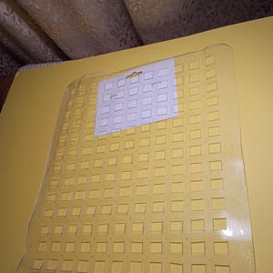 May include: A rectangular, clear plastic mat featuring a grid of square cutouts throughout its surface. A white rectangular paper header card with a central hanging hole is attached to the top section of the clear plastic grid mat.