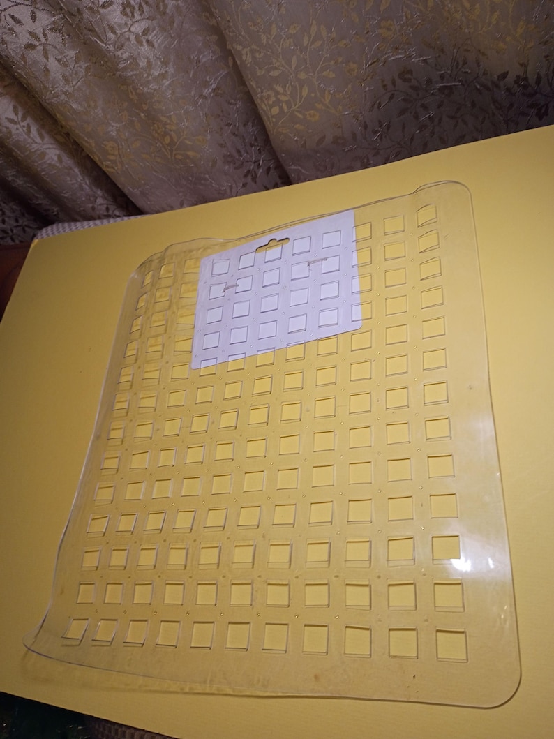May include: A rectangular, clear plastic mat featuring a grid of square cutouts throughout its surface. A white rectangular paper header card with a central hanging hole is attached to the top section of the clear plastic grid mat.