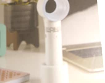May include: A white, handheld personal fan with a circular fan head and a cylindrical handle. The fan is shown on a white surface, with a blurred background including a potted plant and a notebook.