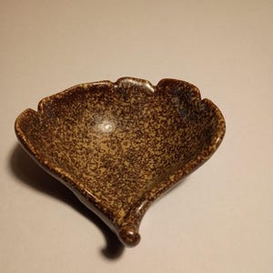 May include: A small, leaf-shaped ceramic dish with a speckled brown and tan glaze. The dish has a scalloped edge and a pointed tip, perfect for holding small items like jewelry or condiments.