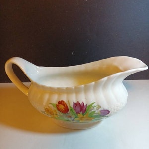May include: A white porcelain gravy boat with a floral design featuring tulips, daffodils, and other flowers in shades of red, yellow, and purple.