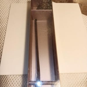 May include: A dark brown, long, slender utensil with a rounded end, presented in a clear rectangular box. The box is encased in a light brown cardboard sleeve. The background features a white surface and a patterned fabric.