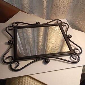 May include: Rectangular black metal framed mirror with decorative scrollwork and small ball accents. The mirror reflects a patterned curtain. The frame has a matte finish and a simple, elegant design, suitable for wall decor.