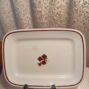 May include: Rectangular white ceramic serving platter with a brown border and a brown floral design. The platter is supported by a dark brown stand. The design features a four-leaf clover.