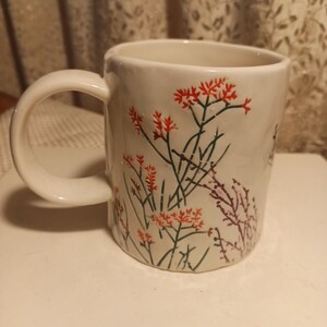 Puede incluir: Taza de cer&aacute;mica blanca con un asa grande, decorada con dise&ntilde;os florales rojos y verdes. La taza tiene forma cil&iacute;ndrica y un asa curva, adecuada para bebidas calientes. El patr&oacute;n floral incluye racimos de flores rojas y tallos verdes.