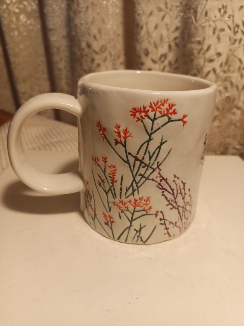 Puede incluir: Taza de cer&aacute;mica blanca con un asa grande, decorada con dise&ntilde;os florales rojos y verdes. La taza tiene forma cil&iacute;ndrica y un asa curva, adecuada para bebidas calientes. El patr&oacute;n floral incluye racimos de flores rojas y tallos verdes.