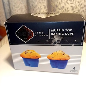 May include: A box of "Dashing Fine Gifts" muffin top baking cups. The box is black and white with the brand name and product description. Two muffins in blue cups are pictured on the front. The box contains 4 cups.