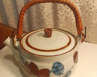 May include: A white ceramic teapot with a brown woven handle and floral pattern. The lid has a brown knob.
