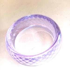 May include: A translucent, light purple bangle bracelet. The bracelet has a geometric, diamond-shaped pattern carved into its surface. The bangle is a solid, circular shape, and appears to be made of glass or a similar material.
