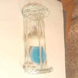 May include: A clear glass hourglass featuring a faceted top and bottom base. The hourglass contains white sand in the upper bulb and bright blue sand in the lower bulb, held together by a central white connector within a transparent frame.