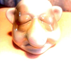 May include: A ceramic figurine of a bald head with closed eyes, a large nose, and small ears. The figurine is primarily beige with brown shading and glossy highlights. The overall design suggests a whimsical or caricature-like aesthetic.