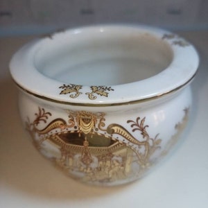 May include: A white ceramic planter with gold trim and a detailed gold design. The planter has a round shape and a wide rim.