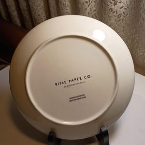 May include: Off-white ceramic plate with the text "RIFLE PAPER CO. for ANTHROPOLOGIE" printed in black. The plate is on a black metal stand. The plate is dishwasher and microwave safe. Made in China.