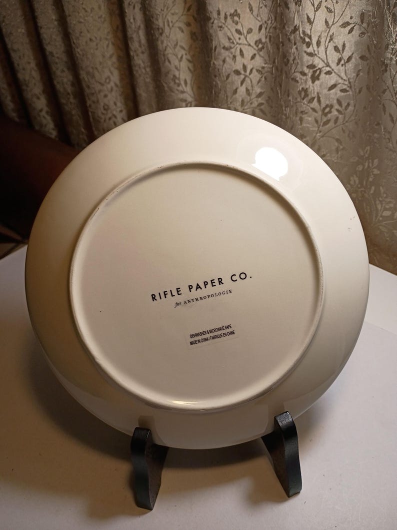 May include: Off-white ceramic plate with the text "RIFLE PAPER CO. for ANTHROPOLOGIE" printed in black. The plate is on a black metal stand. The plate is dishwasher and microwave safe. Made in China.