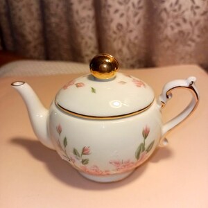 May include: A small, white porcelain teapot with a gold-colored handle and accents. The teapot features a lid with a gold knob and is decorated with pink floral patterns and green leaves. A charming addition to any tea set.