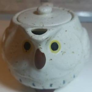 May include: White ceramic teapot shaped like an owl with brown beak and black eyes. The teapot has a lid and a spout.