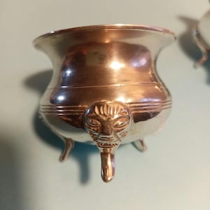 May include: A silver, three-legged, decorative bowl with a face design on the front.