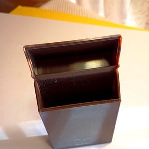 May include: A brown rectangular box with a hinged lid, open to reveal an empty interior. The box has a glossy finish and a small, indistinct logo on the front. The box is made of a hard plastic material.