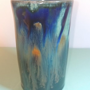 May include: A cylindrical ceramic tumbler featuring a glossy glaze with a drip pattern. The surface displays a blend of deep cobalt blue, teal, and dark green tones, with subtle streaks of amber and light brown running vertically down the body.