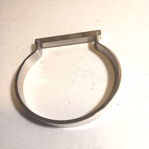 May include: A silver-colored bracelet with a rectangular bar at the top. The bracelet has a simple, minimalist design, with a rounded band that curves around to meet the bar. The metal has a polished finish.