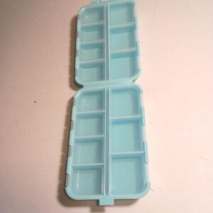 May include: An open, light blue plastic pill organizer with two hinged sections. Each section is divided into multiple compartments of varying sizes. The organizer is designed for storing and organizing pills or small items.