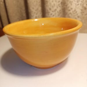 May include: A ceramic bowl with a rounded shape and a warm, yellow-orange color. The bowl has a smooth, glossy finish and a slightly flared rim. It is likely used for serving food or as a decorative item.