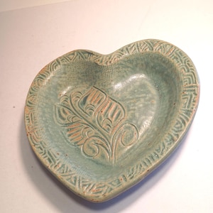 May include: A heart-shaped ceramic dish in a muted teal color. The dish features a carved floral design in the center and a patterned border. The interior has a slightly textured surface, and the edges are rounded.
