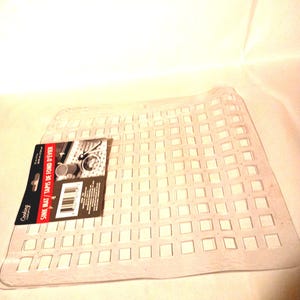 May include: A clear, flexible plastic sink mat featuring a grid of square cutouts. A rectangular product label is attached to the left side, displaying the text "Cooking Concepts" and "SINK MAT / TAPIS DE FOND D'&Eacute;VIER" along with a barcode.