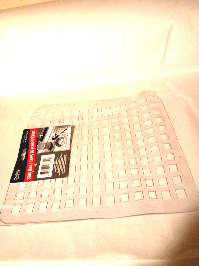 May include: A clear, flexible plastic sink mat featuring a grid of square cutouts. A rectangular product label is attached to the left side, displaying the text "Cooking Concepts" and "SINK MAT / TAPIS DE FOND D'&Eacute;VIER" along with a barcode.