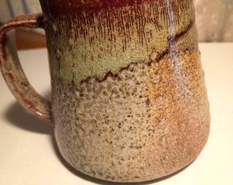 May include: A handmade ceramic mug with a textured surface. The mug features a color gradient, transitioning from a deep red rim to olive green and tan tones. The handle is a matching reddish-brown.