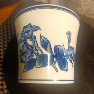 May include: A white ceramic cup with a blue floral design. The design features blue flowers and leaves on a white background.