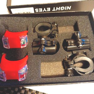 May include: A set of bicycle lights and mounting brackets inside a black foam-lined box labeled NIGHT EYES. The kit includes two red-cased lights with clear lenses and two black adjustable handlebar mounts with tightening knobs and rubber grips.