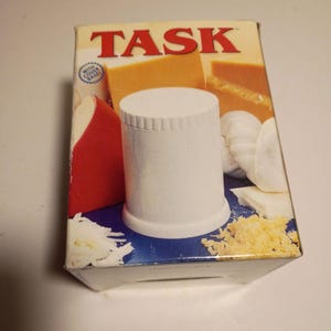 May include: A white cheese grater with a cover base is displayed on a box with the word "TASK" in red. The box features images of cheese and grated cheese. The box is blue and white.