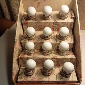 May include: A set of twelve white drawer knobs with a gold-colored base, displayed in a cardboard box with a floral pattern. The knobs are arranged on tiered levels, showcasing their design and presentation.