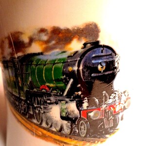 May include: A white ceramic mug featuring a detailed illustration of a vintage green and black steam train. The train has red accents and is emitting brown smoke. The artist's signature, "Derek Bown," is visible near the bottom.