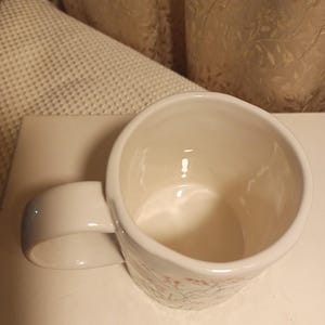 Puede incluir: Taza de cer&aacute;mica blanca con asa redondeada. La taza tiene un dise&ntilde;o floral en rosa y verde. El interior de la taza es blanco. La taza est&aacute; sobre una superficie blanca.