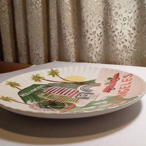 May include: A white ceramic plate with a scalloped edge, featuring a colorful illustration of Hollywood and Los Angeles. The design includes palm trees, a red car, and the words "Hollywood" and "Angeles".