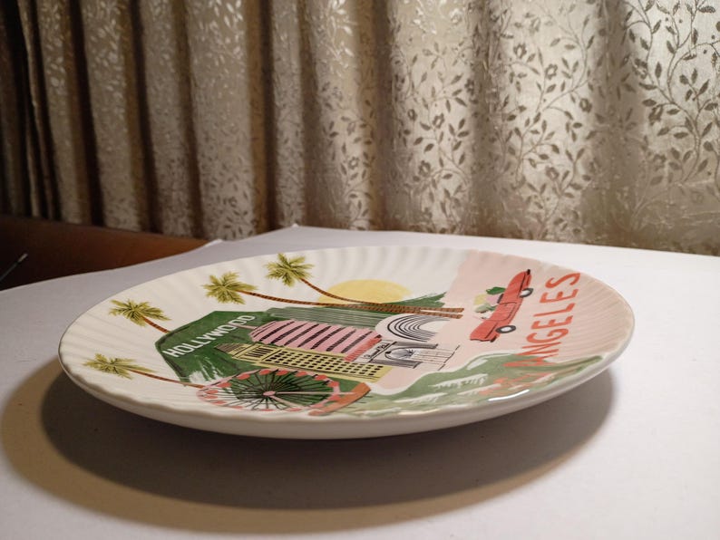May include: A white ceramic plate with a scalloped edge, featuring a colorful illustration of Hollywood and Los Angeles. The design includes palm trees, a red car, and the words "Hollywood" and "Angeles".