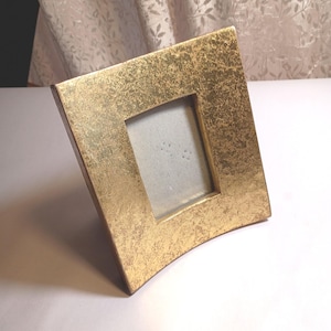 May include: A rectangular picture frame with a metallic gold finish and a distressed, textured surface. The frame features a slightly curved, concave profile and a central rectangular opening for a photograph, set against a plain light background.