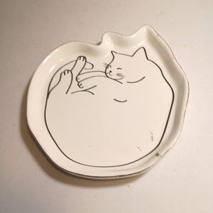 May include: A white ceramic trinket dish shaped like a cat's head with a black line drawing of a curled-up cat in the center. The dish features a smooth, glossy finish and a raised edge that follows the outline of the cat's ears and head.