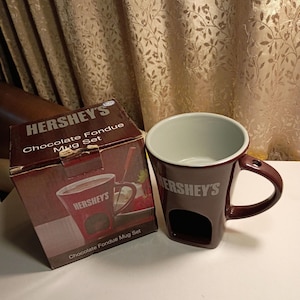 May include: A Hershey's Chocolate Fondue Mug Set. The mug is dark brown with a white interior and a handle. The mug has a rectangular opening on the side. The box is brown with the Hershey's logo and product information.