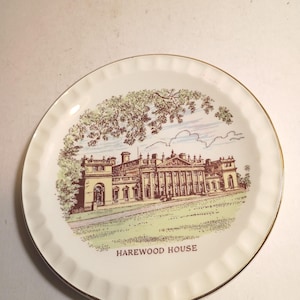 May include: A round, cream-colored ceramic plate with a scalloped edge and a gold-tone rim. The center features a detailed illustration of Harewood House with trees and a lawn, with the text HAREWOOD HOUSE printed in brown below the building.