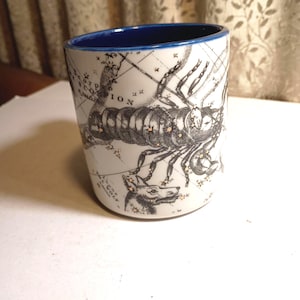 Peut inclure: Mug en c&eacute;ramique blanche avec un bord int&eacute;rieur bleu. Le mug pr&eacute;sente une illustration en noir et blanc d'un scorpion et un motif de carte. Le mot "SCORPIO" est visible sur le mug.