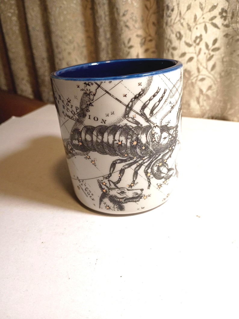 Peut inclure: Mug en c&eacute;ramique blanche avec un bord int&eacute;rieur bleu. Le mug pr&eacute;sente une illustration en noir et blanc d'un scorpion et un motif de carte. Le mot "SCORPIO" est visible sur le mug.