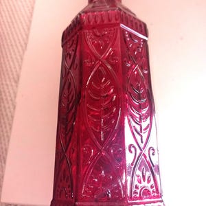 May include: A ruby red glass bottle with an ornate, embossed design. The bottle has a rectangular shape with a series of leaf-like patterns and decorative flourishes. The bottle's surface is smooth and reflective, with a slightly textured base.
