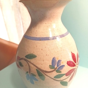 May include: A beige ceramic vase with a floral design. The vase has a wide mouth and a rounded body. The floral design features blue and red flowers with green leaves.