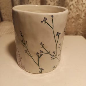 Puede incluir: Una taza de cer&aacute;mica blanca con una superficie ligeramente texturizada. Delicados tallos verdes y peque&ntilde;as flores moradas est&aacute;n pintados a mano en el exterior. La taza tiene forma cil&iacute;ndrica y un borde liso.